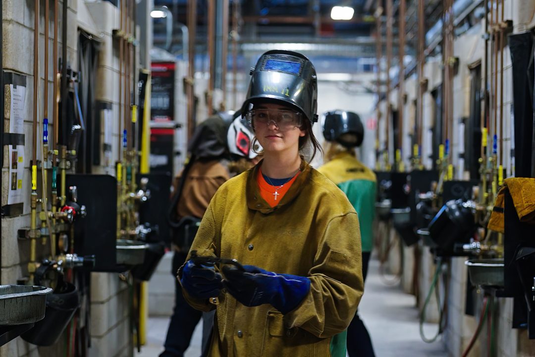JOATWelding(5of14) A yound woman wearing a welders mask and welding gear stands at an ARC station
